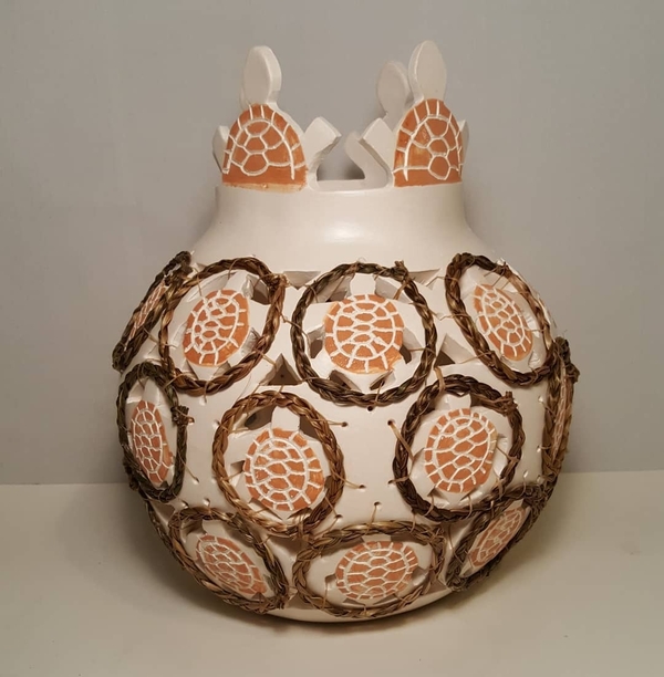A ceramic pot with intricate designs and patterns. The pot is white and features circular motifs resembling turtle shells, highlighted in white and earth-red hues. The circles are surrounded by woven sweetgrass, and the top has unique cut-out shapes of turtles.
